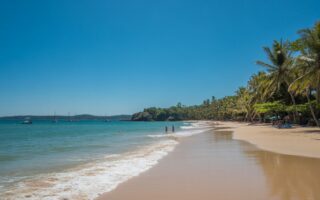 Praia com areia branca, mar azul e palmeiras ao fundo sob céu claro.