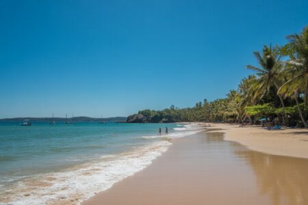 Praia com areia branca, mar azul e palmeiras ao fundo sob céu claro.