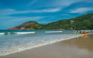 Praia com areia clara, água azul transparente, colinas verdes e algumas pessoas surfando em ondas pequenas.