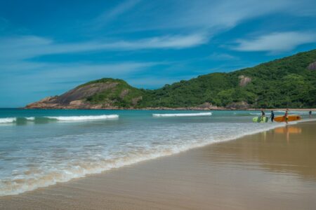 Praia com areia clara, água azul transparente, colinas verdes e algumas pessoas surfando em ondas pequenas.