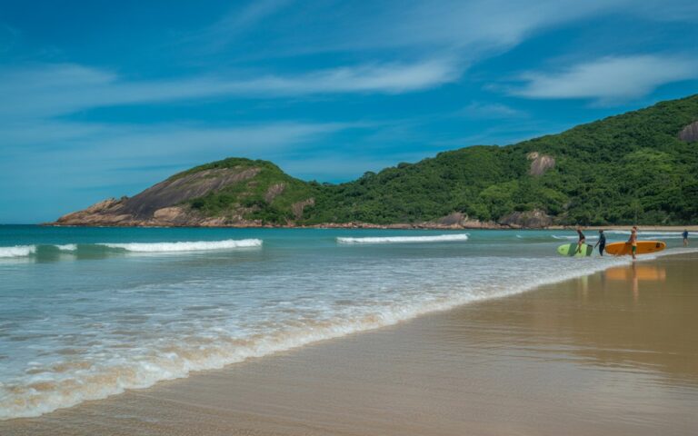 Praia com areia clara, água azul transparente, colinas verdes e algumas pessoas surfando em ondas pequenas.