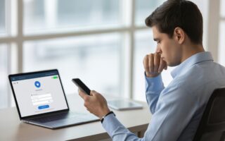 Homem jovem sentado em uma mesa de escritório olhando para um smartphone, com um laptop aberto mostrando uma carteira digital.