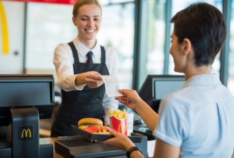 Cliente recebendo um recibo de um caixa sorridente dentro de um restaurante McDonald's moderno, com alimentos típicos da rede sobre a bandeja.