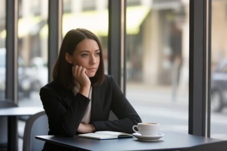 Mulher pensativa sentada sozinha em uma mesa de café moderna perto de uma janela grande, olhando para longe com expressão reflexiva.