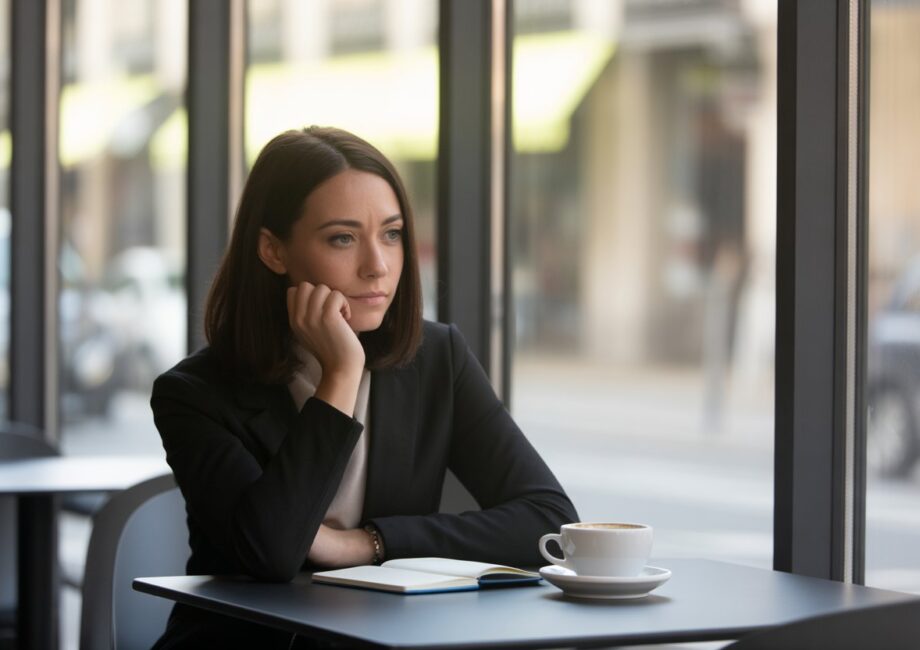 Mulher pensativa sentada sozinha em uma mesa de café moderna perto de uma janela grande, olhando para longe com expressão reflexiva.