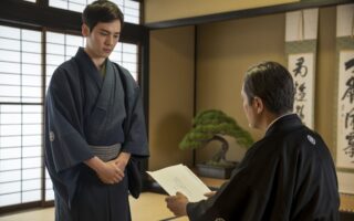 Um jovem de kimono escuro está em um quarto tradicional japonês, conversando seriamente com um homem mais velho sentado, transmitindo uma atmosfera de disciplina e respeito.