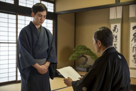 Um jovem de kimono escuro está em um quarto tradicional japonês, conversando seriamente com um homem mais velho sentado, transmitindo uma atmosfera de disciplina e respeito.