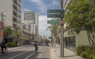 Rua moderna em um bairro brasileiro com prédios residenciais e comerciais, pessoas caminhando e árvores ao redor.