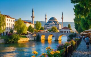 Vista da cidade de Prizren com a ponte sobre o rio, edifícios históricos, mesquita ao fundo e pessoas caminhando nas ruas.