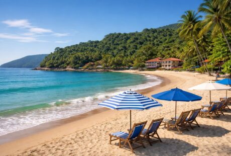Praia de Ubatuba com cadeiras de praia, guarda-sóis, vegetação tropical e hotéis à beira-mar sob céu azul.