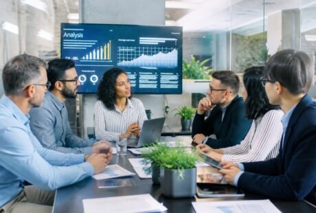 Grupo de pessoas em um escritório moderno discutindo em volta de uma mesa com laptops e gráficos em uma tela ao fundo.