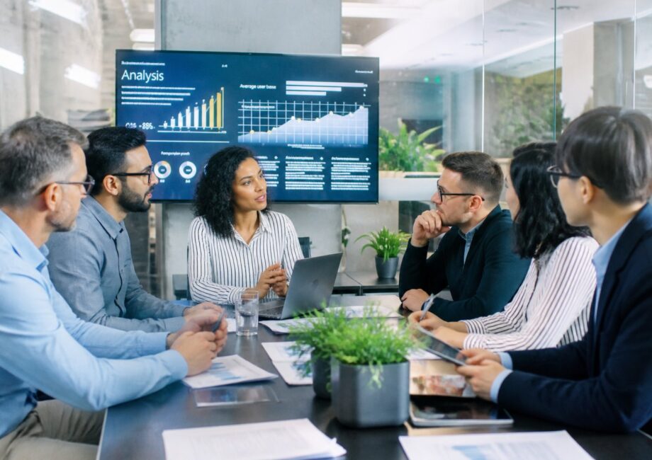 Grupo de pessoas em um escritório moderno discutindo em volta de uma mesa com laptops e gráficos em uma tela ao fundo.