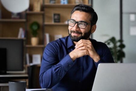 Homem de meia-idade sentado em um escritório moderno, sorrindo e olhando para o lado, com estantes de livros e plantas ao fundo.