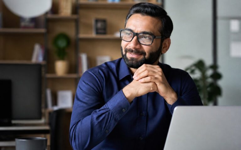 Homem de meia-idade sentado em um escritório moderno, sorrindo e olhando para o lado, com estantes de livros e plantas ao fundo.