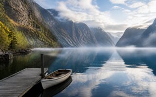 Paisagem de um fiorde na Noruega com penhascos rochosos, árvores verdes, água calma e um barco vazio amarrado a um pequeno cais de madeira.