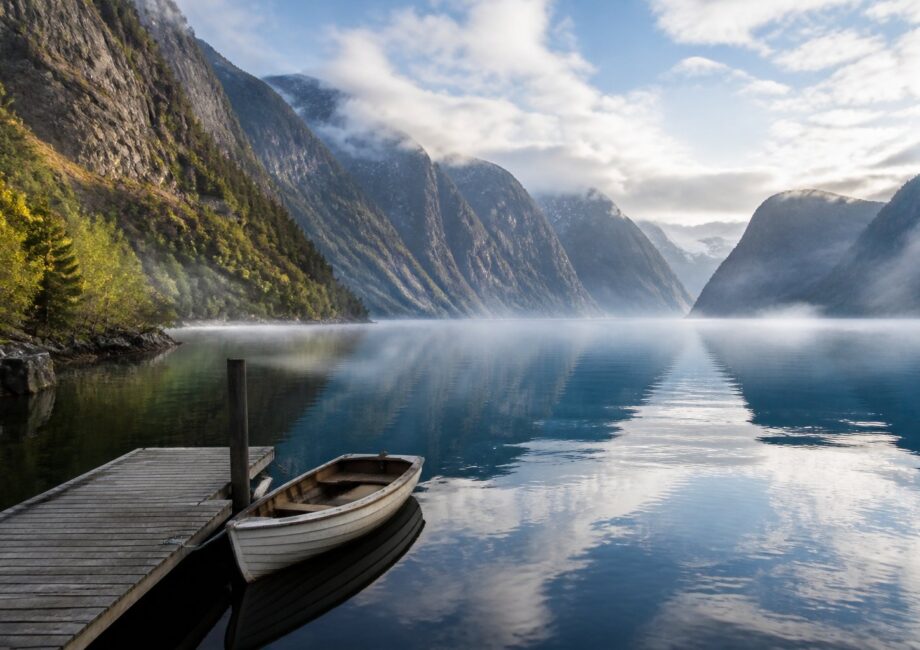 Paisagem de um fiorde na Noruega com penhascos rochosos, árvores verdes, água calma e um barco vazio amarrado a um pequeno cais de madeira.